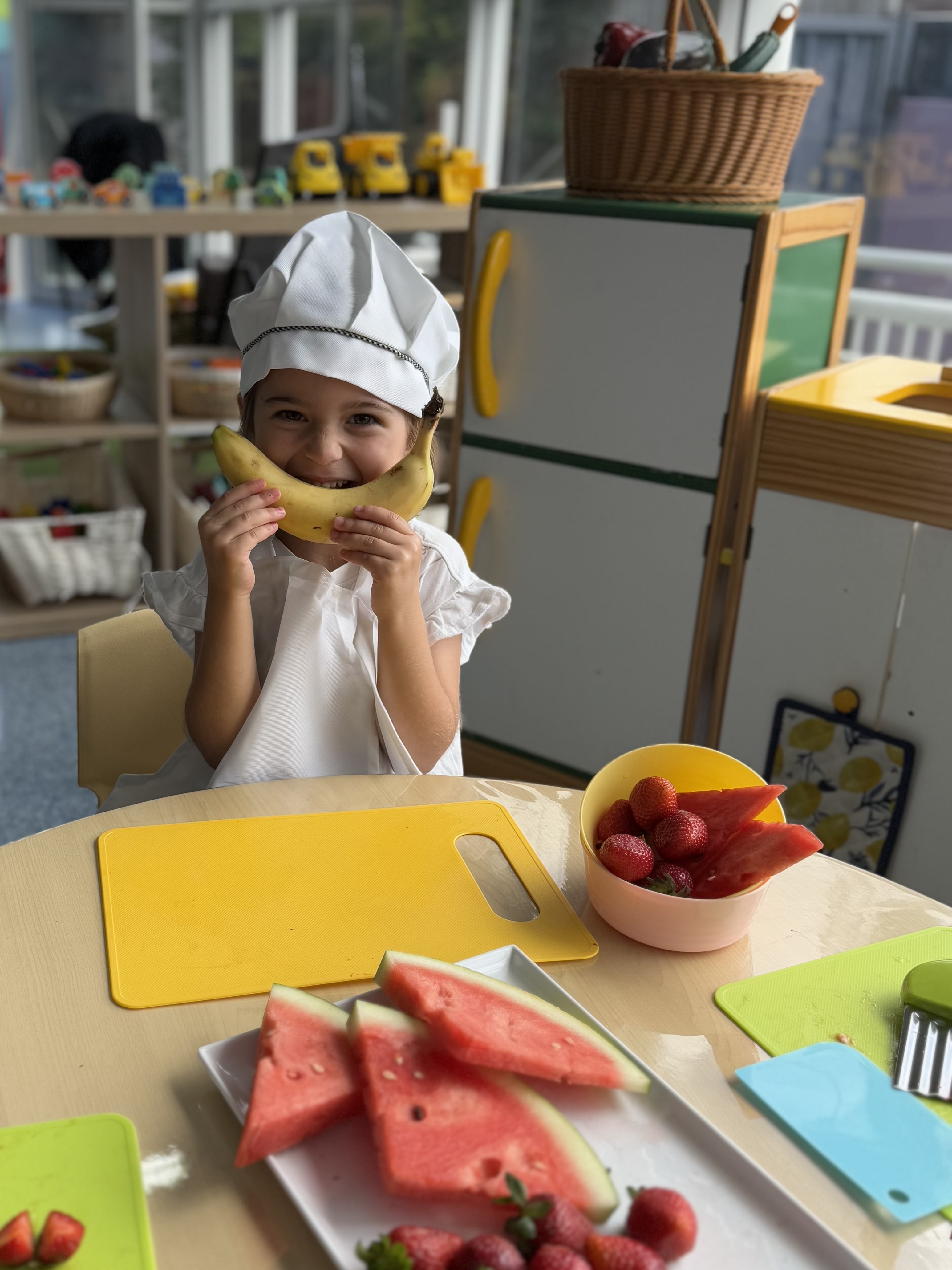 Smiling child dressed as a chef with a banana in front of her face, ready to enjoy a snack.